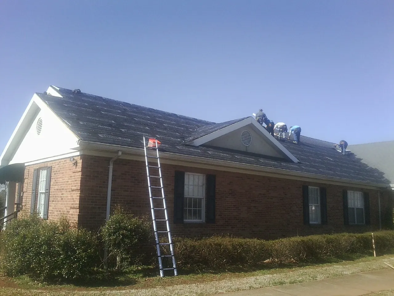 Crew tearing shingles off a brick ranch roof with a ladder set — Metal Roof Repair work by our crew in South Sutton